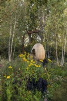 Yeo Valley Organic Garden. A suspended, steam-bent, oak egg seat which forms the focal point of the garden. Naturalistic perennial meadow with burnt wood sculptures in the foreground. Planting includes silver birches, Betula pendula, Kniphofia 'Tawny King', Rudbeckia laciniata 'Herbstonne', Astrantia major var. rosea, Calamagrostis brachytricha, and various Persicarias.