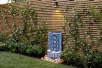 Slatted fence with trained Trachelospermum jasminoides and arabic-style water feature with spotlight mounted on fence