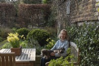 Women sitting at the garden table with her dogs, surrounded by Narcissus 'Hawera', Trachelospermum jasminoides, with Buxus sempervirens spheres and a Beech hedge - Fagus sylvatica in the background.