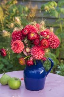 Bouquet of orange Dahlias and Helichrysums with Nigella seedheads in dark blue pottery jug with apples on table