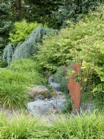 Metal sculpture of heron and bullrushes in dry stream bed featuring foliage of daylilies, spike winterhazel and weeping blue atlas cedar