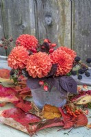 Orange Dahlias arranged with rosehips and blackberries and purple foliage in metal pot against wooden background
