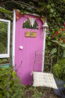 A recycled door, painted bright pink and with handle and fun name plate attached gives colour and interest to a dark fence panel at the end of a cottage style garden with an old metal chair beside it providing a place to sit and contemplate. 