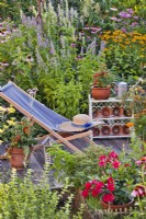 Decked patio with deck chair and pot grown herbs, vegetables and flower surrounded with herbaceous flowerbed.