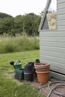 Stacked plastic pots and watering equipment outside garden shed with wildflower meadow beyond. July. Summer.