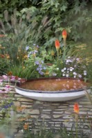 Circular pool of water atop a small wall bordering a garden.  With Campanula lactiflora, Erigeron karvinskianus, Kniphofia grandiflora and Pennisetum.