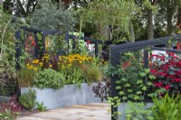 A raised bed is filled with hot coloured flowers to attract pollinators: rudbeckias, crocosmias and dahlias, offset by dark canna leaves and ornamental grasses.