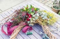 Wreath, secateurs, wire, ribbon, moss, ivy, pussy willow, skimmia japonica, hebe and carnations laid out on a table