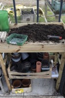 A potting bench ready for sowing seeds with a seed tray and trowel. Gloves and plant pots of varying sizes are beneath the potting bench.