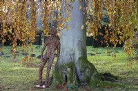 A woven willow figurine by the sculptor Paul Baines leans against Fagus sylvatica pendula, weeping beech.