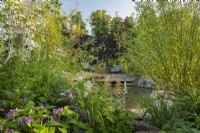 View across pond to wooden deck with seating area. RHS Garden For A Green Future. Designed by Jamie Butterworth. RHS Hampton Court Palace Garden Festival Show 2021, July 2021