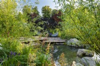 View across pond to seating area on wooden deck. RHS Garden For A Green Future. Designed by Jamie Butterworth. RHS Hampton Court Palace Garden Festival Show, July 2021