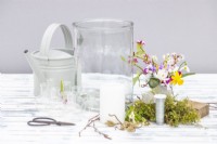 Large glass container, small glass bottles, birch twigs, corkscrew hazel, moss, candle, flower snips, wire, wooden block, watering can and various small flowers laid out on a wooden surface