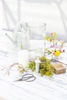 Large glass container, small glass bottles, birch twigs, corkscrew hazel, moss, candle, flower snips, wire, wooden block, watering can and various small flowers laid out on a wooden surface