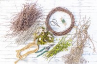 Wreath, pruning scissors, wire, secateurs, rope, moss, feathers, birch twigs, hazel twigs, hawthorn twigs, russian sage and Dried seed heads laid out on a wooden surface