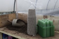A bulk load of green plastic seed trays and grey plant pots sit on a potting bench beside a pile of compost and a wooden handled scoop in a small commercial nursery. 