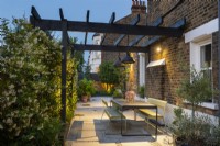 Illuminated dining area in long narrow space with table and chairs under pergola next to the wall of scented Trachelospermum jasminoides. 
