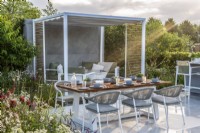 Morning sun on patio with dining table and metal pergola with seating under.  Lower Barn Farm: The Bounce Back Garden, RHS Hampton Court Palace Garden Festival 2021