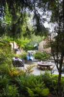 View across woodland garden with shade tolerant plants to sunken seating area with freestanding chairs and fire pit.