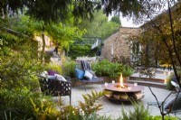 View across beds with shade tolerant plants to sunken seating area with freestanding chairs and fire pit.