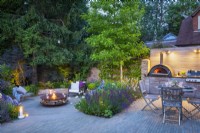 Illuminated sunken garden and outdoor dining area with wooden table, chairs and bespoke kitchen unit with integrated pizza oven and bbq grill at night.