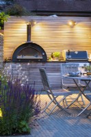 Illuminated outdoor dining area with wooden table, chairs and bespoke kitchen unit with integrated pizza oven and bbq grill at night. 