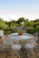Rustic wire table and chairs on a patio with terracotta pot planted with Verbena, greenhouse and rose garden beyond.   The Old Rectory, Isle of Wight