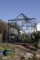 A new greenhouse constructed on a vegetable patch. A water butt sits to the left of the greenhouse and there are various pots in the foreground. 