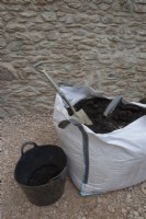 A bulk quantity of compost in a dumpy bag sits beside a plastic trug, half filled with compost with a spade in the dumpy bag. Both sit in front of a newly constructed wall. 