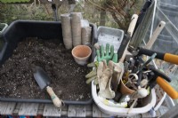 A potting bench in a greenhouse sits beside a tub holding various gardening tools including mallet, shears, scissors and several pairs of gardening gloves. A packet of eco friendly pots sits on the potting bench. 