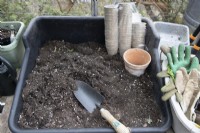 A potting bench in a greenhouse is ready with compost, eco friendly plant pots and a trowel. Gardening gloves are stored at the side. 
