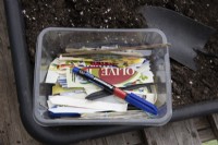 A box of plant labels with a permanent marker sit in a potting tray on compost beside a trowel. The labels are made from cut up plastic tubs such as margarine tubs. 