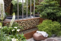 A small contemporary garden incorporates a jacuzzi with a sunken area housing a fire pit with seating and a water feature. It is enclosed in maples, tree ferns, palms, a wheel tree and white flowers.