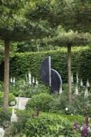 Framed by flat topped hawthorn trees, a sculpture of steel and slate, titled 'Connected' by Tom Stogdon creates a focal point in The Perennial Garden 'With Love', it is surrounded by digitalis purpurea f. albiflora - Designer: Richard Miers - Sponsor: Perennial