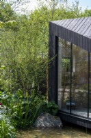 A charred larch cabin overlooks a pond surrounded by Betula nigra -  A Garden Sanctuary by Hamptons,  RHS Chelsea Flower Show 2022