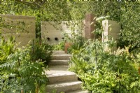 Steps lead up through lush green planting to a pipe water feature and bench - The Mind Garden, RHS Chelsea Flower Show 2022 - Gold Medal