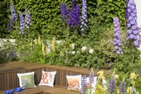 Sunken seating area with wooden bench and cushions planted above with delphiniums, lupins and irises - New Blue Peter Soil Garden. 