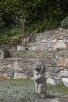 Terracing is broken up by a stone seat with home-made mosaic snake on the back of it. An olive tree in a pot sits on a terrace and a stone lion on a lower terrace. Briar Cottage Garden. Devon NGS garden. Spring