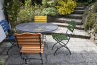A round glass table sits on a patio area with four different coloured seats. Briar Cottage Garden. Devon NGS garden. Spring