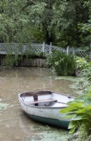 A wooden rowing boat on a pond with a bridge in the background. Lewis Cottage, NGS Devon garden. Spring.