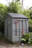 A wooden shed labelled with a sign for recycling and rubbish bins has old, long-handled, wooden tools hung on the side of it. Lewis Cottage, NGS Devon garden. Spring.