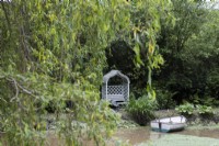 A wooden rowing boat on a pond with a wooden arbour seat on the edge and a pair of waders beside the seat with willow tree foliage in foreground. Lewis Cottage, NGS Devon garden. Spring.