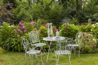 Seating area at the hydrangea bed with garden hydrangeas, crested lilies, Hydrangea quercifolia 'Snowflake'. Lifestyle furniture metal painted white table chairs lawn.
