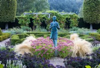 The Formal Garden, with a knot garden  of Buxus sempervirens, Box and Berberis thunbergii atropurpureum, Purple Barberry. The statue of a young girl  stands amongst Sedum 'Autumn Joy', Heliotropium arborescens, Heliotrope, and Stipa tenuissima, Mexican Feather Grass.  The garden is walled in by a tall Taxus baccata,  Yew hedging.  Waterperry Gardens, Wheatley, Oxfordshire, UK