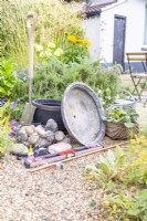 Shallow container, faux barrel container, cobbles, water pump, glue, spirit level, copper pipe, pipe cutter, pencil, spade, watering can and plants laid out on the ground