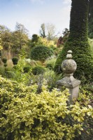 Stone finial topped with pebbles in a formal country garden in November, amongst evergreen shrubs including euonymus, clipped yew and box.