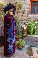 Venetian mannekin dressed in velvet robe and jewelled and feathered headress in enclosed courtyard with various plants in pots including arum lilies and heucherella.