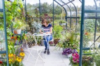 Woman sitting at table writing in a greenhouse filled with various plants and containers