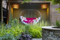 Illuminated outdoor reading room with hanging wooden bench next to the borders with purple and lime Heuchera. 