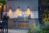 Illuminated outdoor kitchen with ornamental tiles, grey wall, bbq and storage.  